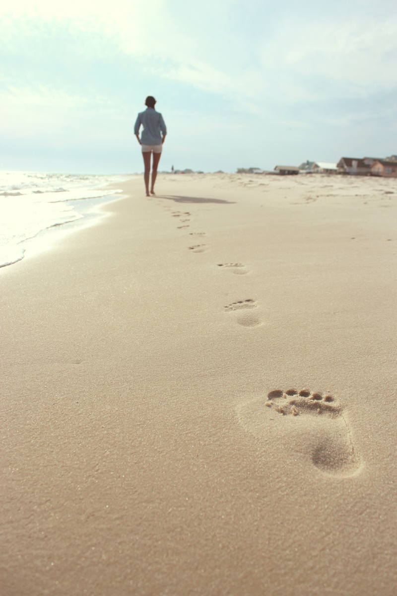 Footsteps on a beach with a young woman walking in the distance.
